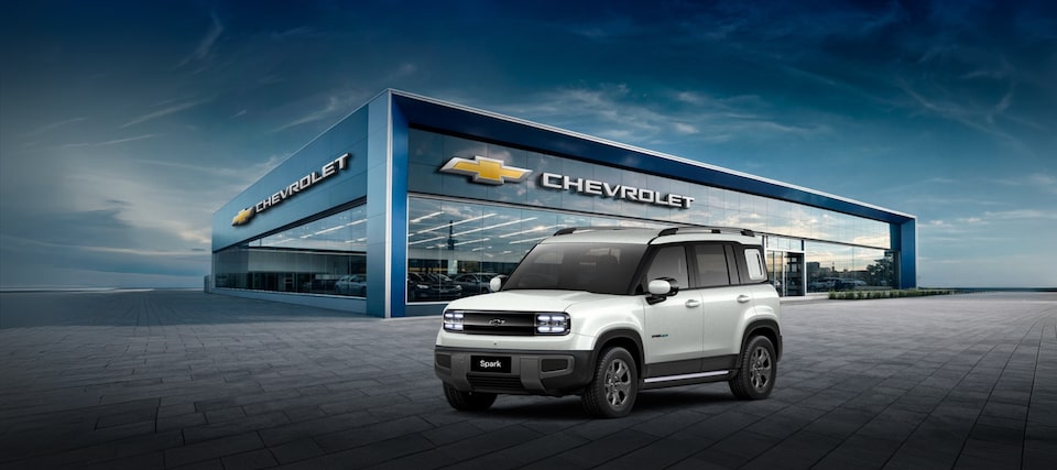 Um SUV elétrico Spark EUV branco está estacionado em frente a uma concessionária Chevrolet moderna sob um céu parcialmente nublado.