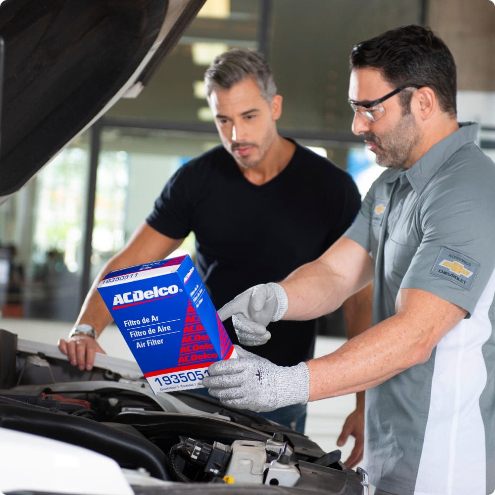 Dois homens na frente de um carro com o capô aberto. O mecânico está mostrando uma caixa de filtro de ar para o dono do carro.