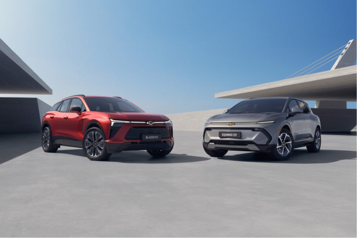 em uma área aberta, vemos um Blazer EV vermelho e um Equinox EV cinza, ambos em um ângulo em que vemos a frente e a lateral dos veículos