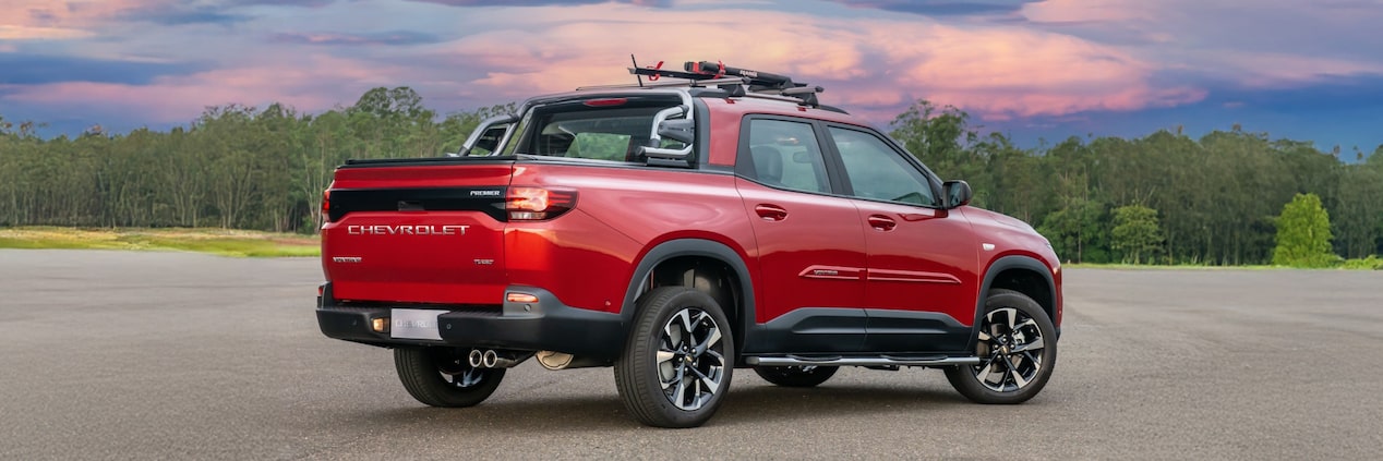 Chevrolet Montana vermelha em imagem ambientada, estacionada a céu aberto, em posição 3/4 de costas.