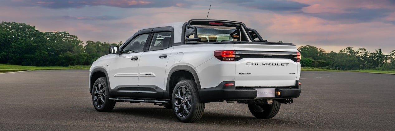 Chevrolet Montana branca em imagem ambientada, estacionada a céu aberto, em posição 3/4 de costas.