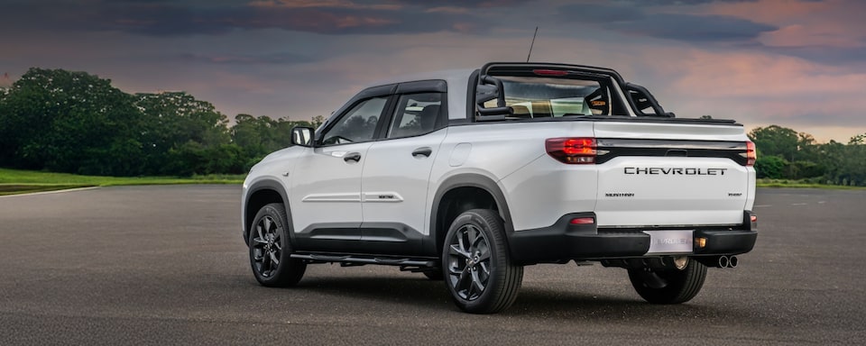 Chevrolet Montana branca em imagem ambientada, estacionada a céu aberto, em posição 3/4 de costas.