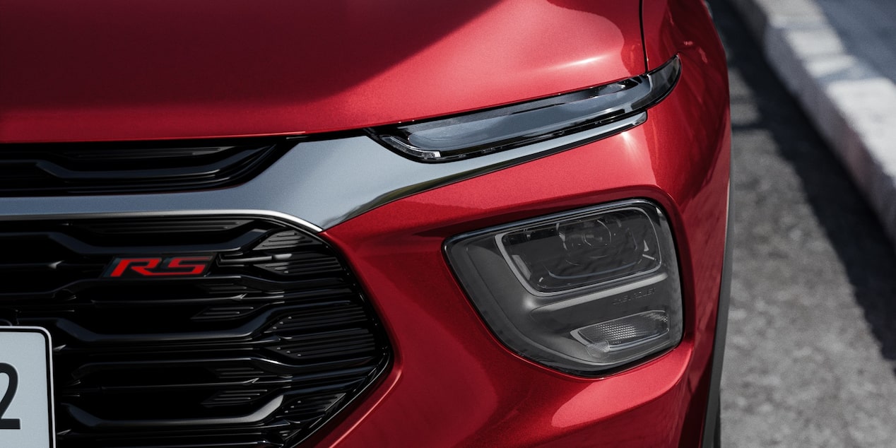 Close do conjunto óptico dianteiro da Chevrolet Montana na cor Vermelho Scarlet com farol de LED e emblema RS em fundo urbano