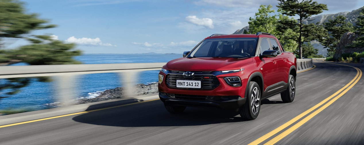 Chevrolet Montana versão RS na cor Vermelho Scarlet em rua asfaltada à beira-mar, com guarda-corpo branco, árvores e céu azul