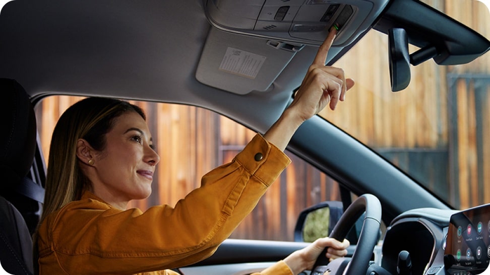Mulher tocando botão do OnStar no teto da Chevrolet Montana, vista interna com acabamento escurecido e cenário de madeira ao fundo