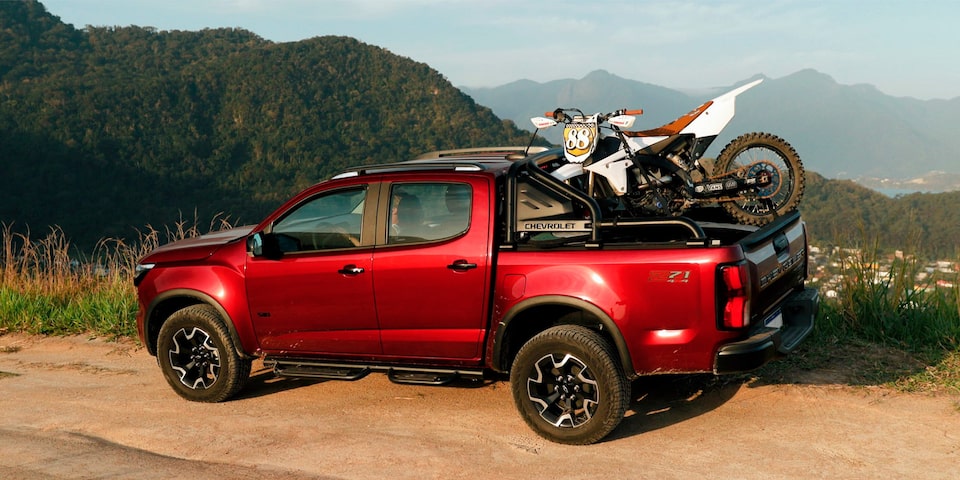 Chevrolet S10 Z71 2026 vermelha usada pelo o influenciador Lucas Xaparral estacionada em local de terra batida à beira-mar. A caçamba está carregada com uma prancha de surfe e uma motocicleta.