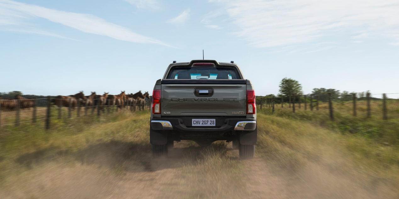 Chevrolet S10 cinza vista pela traseira, em movimento por uma estrada de terra, com cavalos e cercas ao fundo.