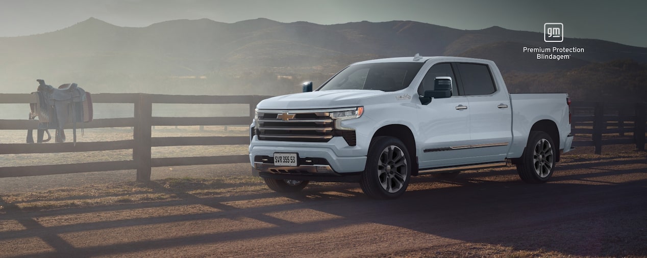 Picape Chevrolet Silverado 2026 branca quartzo estacionada em área rural, com cerca de madeira, cavalo ao fundo e montanhas ao entardecer. O texto destaca “Silverado 2026” e “GM Premium Protection Blindagem”.