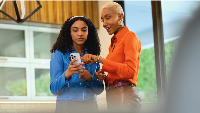 Dupla de pessoas em ambiente interno, uma usando camisa azul e outra camisa laranja, olhando juntas para a tela de um celular.