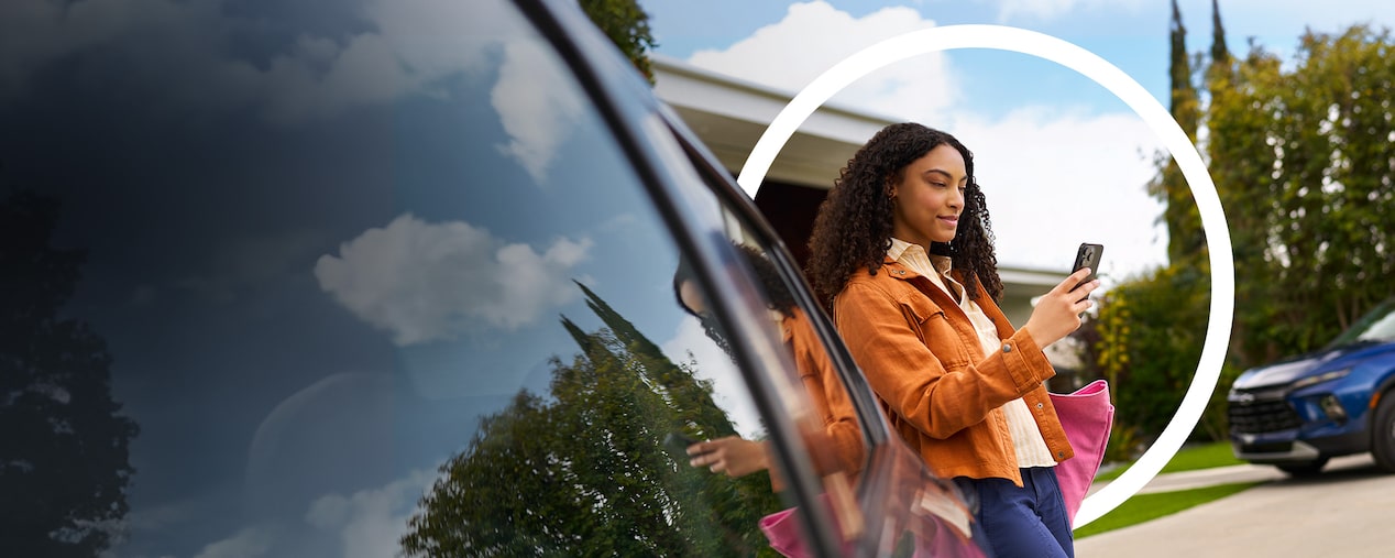 Uma mulher negra está apoiada em um carro enquanto usa o smartphone. Ela usa uma jaqueta laranja e uma bolsa rosa. Um círculo, usado como grafismo de OnStar, está em volta dela.