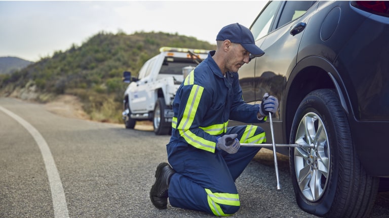 Um profissional de assistência mecânica, uniformizado e usando colete refletivo, troca o pneu de um carro na estrada, com um caminhão de apoio estacionado ao fundo.