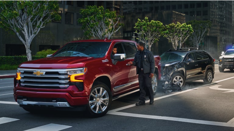 Picape Chevrolet vermelha parada após colisão em via urbana, com um homem em pé próximo ao veículo e outro carro danificado ao fundo.