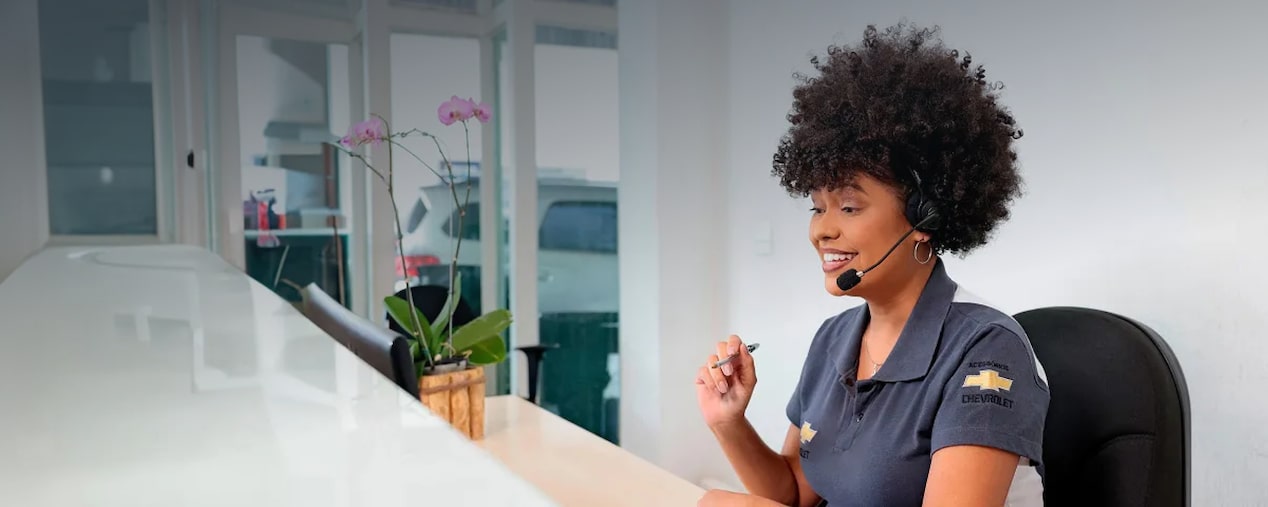 Uma mulher trabalhando em frente ao computador, ela usa um fone com microfone. Em sua camiseta, conseguimos ver o logo da Chevrolet na manga.