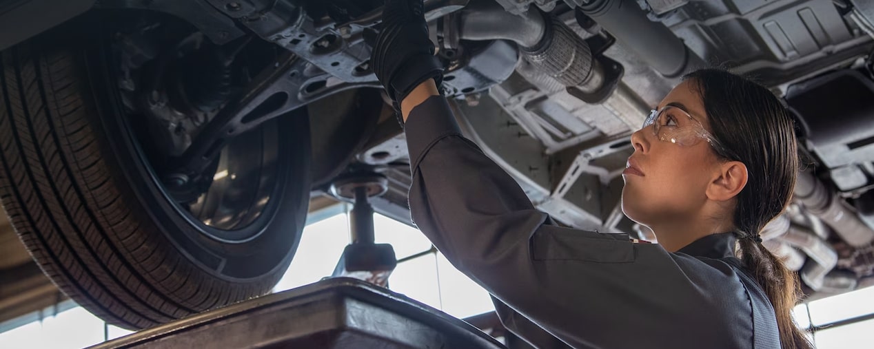 Uma mecânica usando macacão e  óculos de proteção está trabalhando na parte de baixo de um carro.