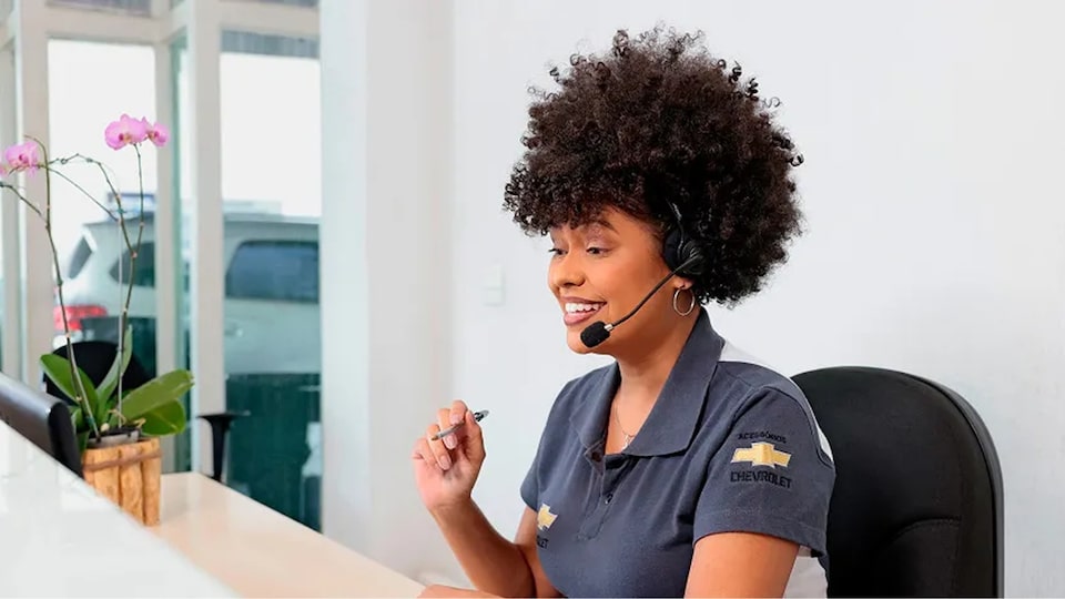 Mulher negra em primeiro plano, envergando uma camisa polo com o logo Chevrolet na manga e usando um head phone para atender ligações. Em segundo plano, um Chevrolet estacionado.