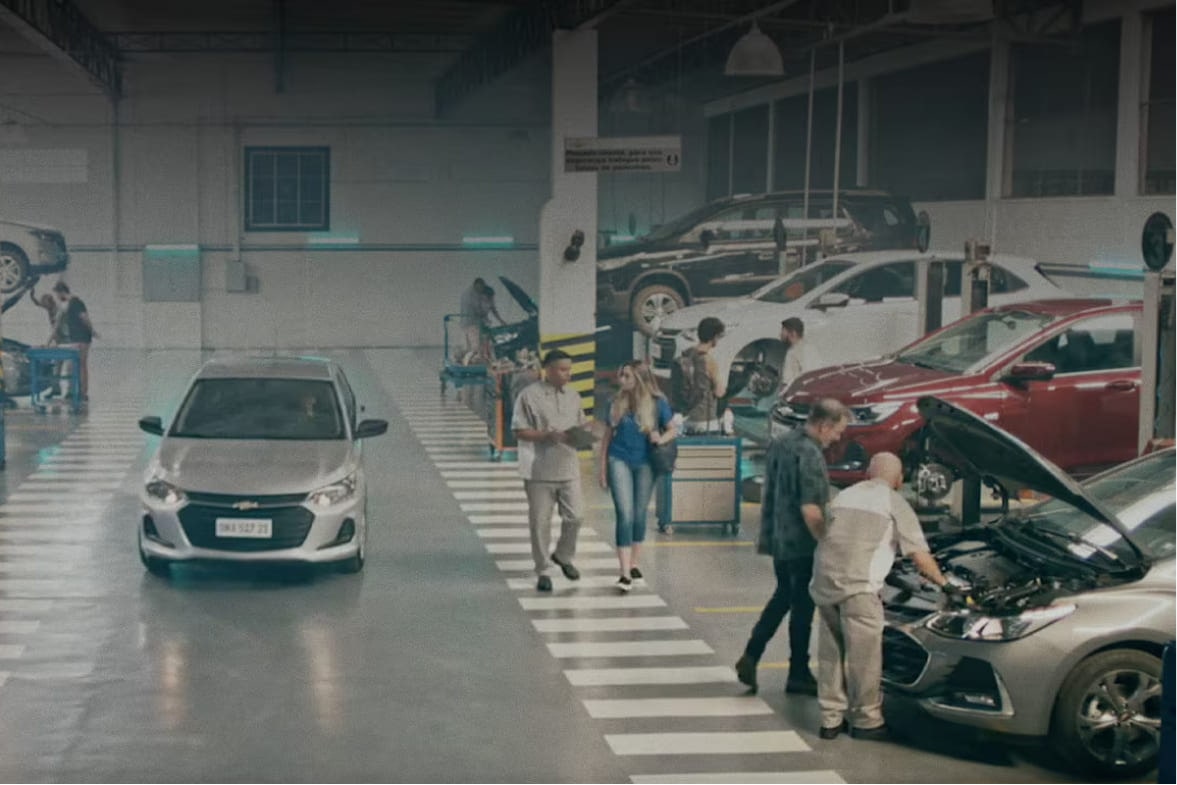uma concessionária Chevrolet com vários carros sendo trabalhados. Um está com o capô levantado. Ao fundo, outro está levantado e no meio da imagem um novo veículo está chegando ao local. Também vemos mecânicos trabalhando.
