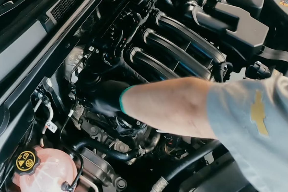 vemos um capô aberto com destaque para o motor. Um braço de um mecânica está trocando uma peça. Vemos o logo da Chevrolet na manga da camisa.