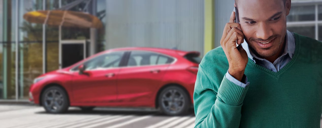 em primeiro plano, vemos um homem usando uma camisa quadriculada e um suéter verde enquanto fala no smartphone. Em segundo plano, um Chevrolet vermelho.