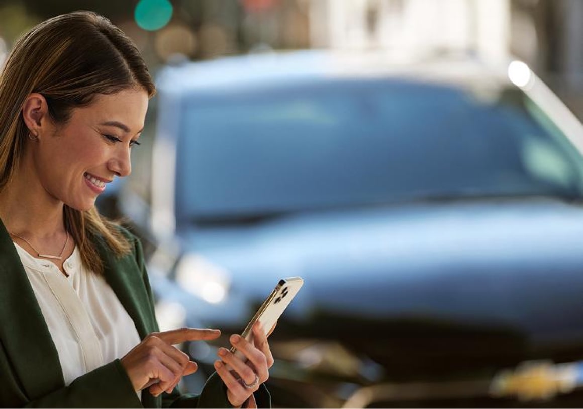 em primeiro plano, vemos uma mulher de camisa branca e blazer verde, soliciatando o Chevrolet Road Service no smartphone. Em segundo plano, vemos um Chevrolet preto.